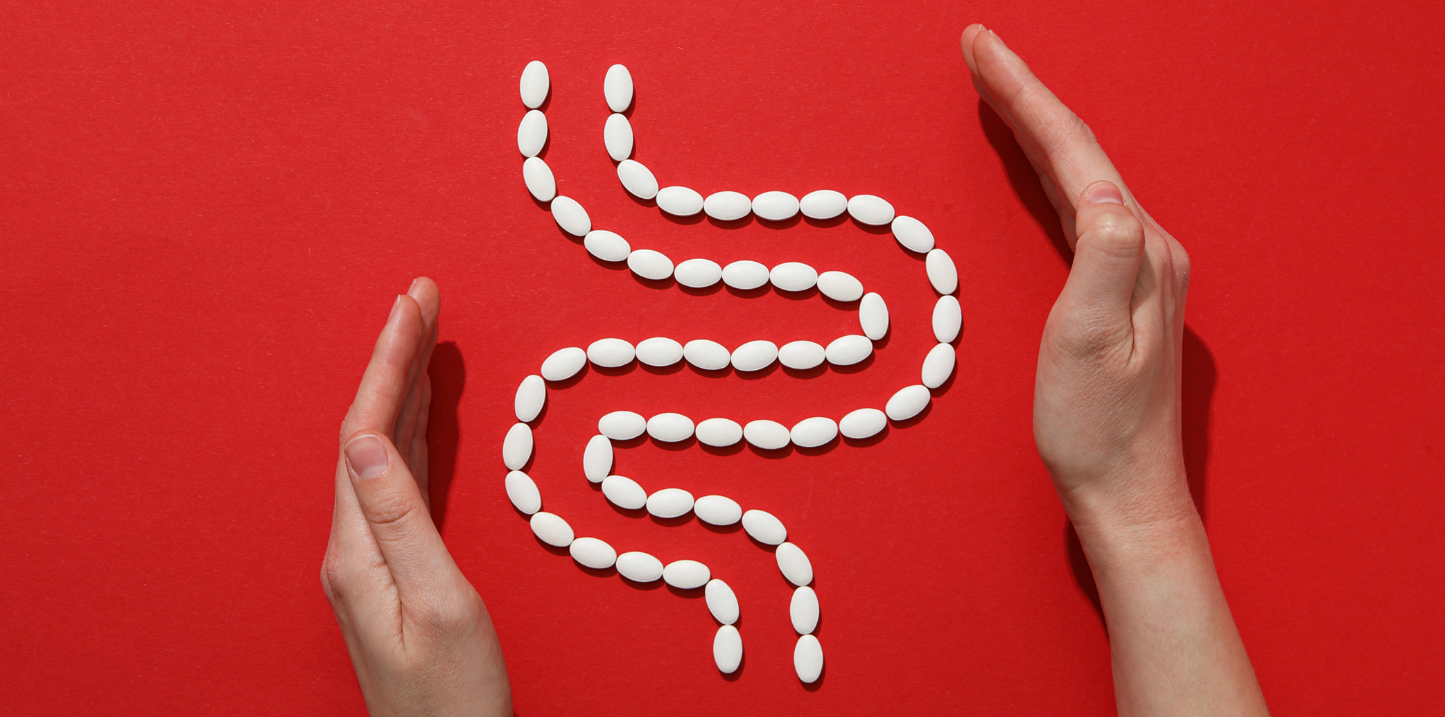 Two hands surrounding a stylized digestive tract made of white oval pills on a red background, symbolizing gut health and digestive care.
